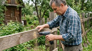 Мужчина сверлит аккумуляторным шуруповёртом доску забора на дачном участке