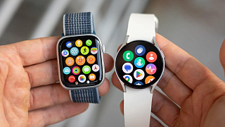 Apple Watch и Samsung Galaxy Watch в двух руках рядом, на обоих экранах открыто меню приложений
