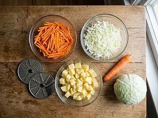 Морковь соломкой, шинкованная капуста и кубики картофеля в мисках, рядом два режущих диска комбайна