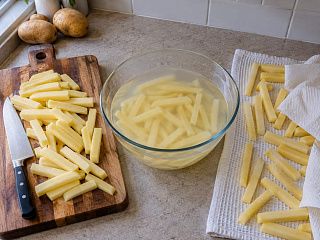 Сырые бруски картофеля одной толщины: на доске, в миске с холодной водой и на полотенце для обсушивания